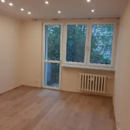 Pusty pokój po remoncie z jasnymi panelami podłogowymi, świeżo pomalowanymi beżowymi ścianami, białym grzejnikiem i oknem balkonowym z widokiem na zieleń.