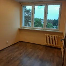 Pusty pok&oacute;j z drewnianą podłogą, pomarańczowymi ścianami i białym grzejnikiem pod oknem z widokiem na zieleń.