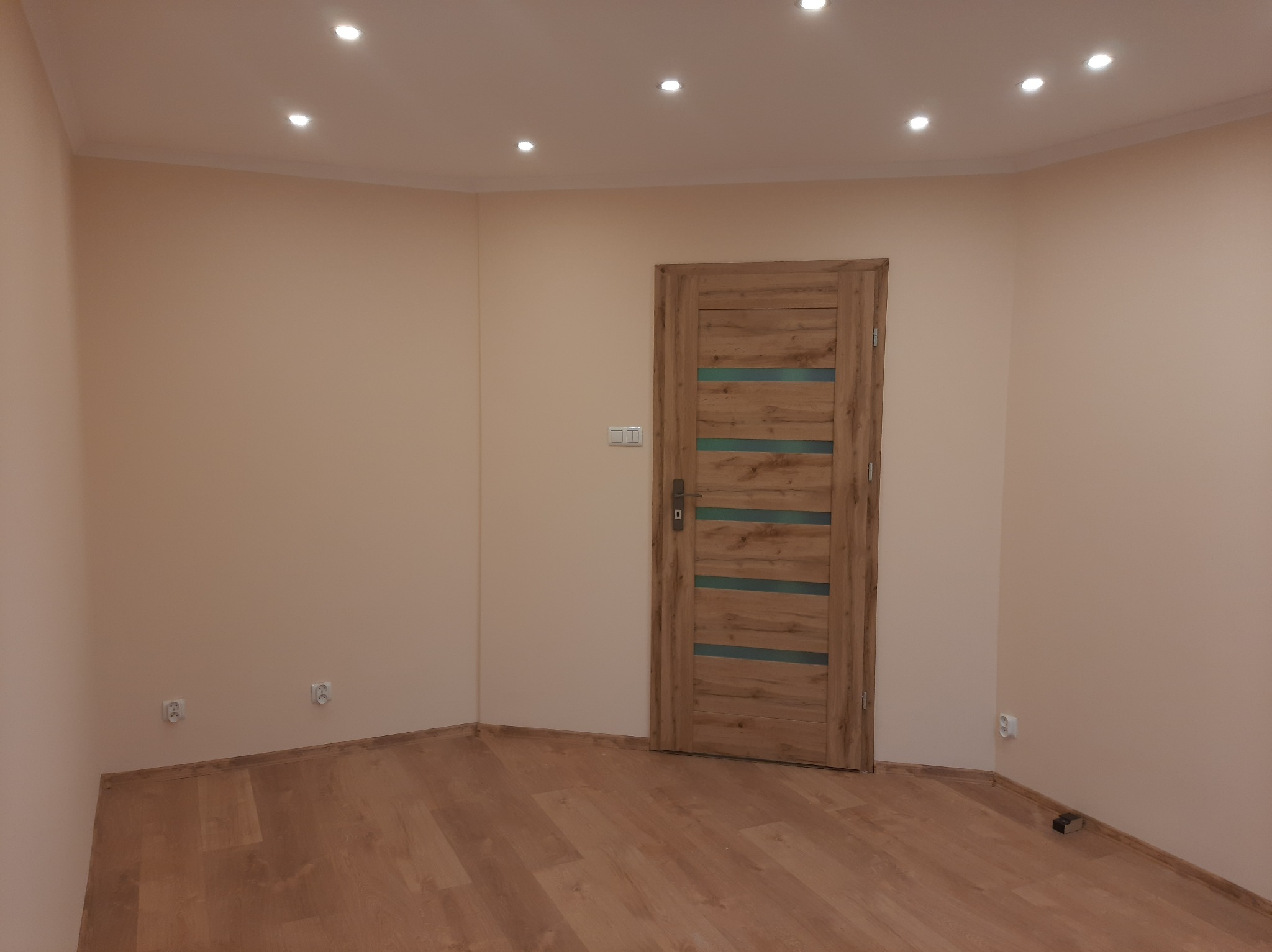 Wnętrze pokoju po remoncie: ściany pomalowane na jasny beż, drewnopodobna podłoga, zamontowane drzwi z drewnianą okleiną i poziomymi, przezroczystymi paskami, oświetlenie sufitowe punktowe.