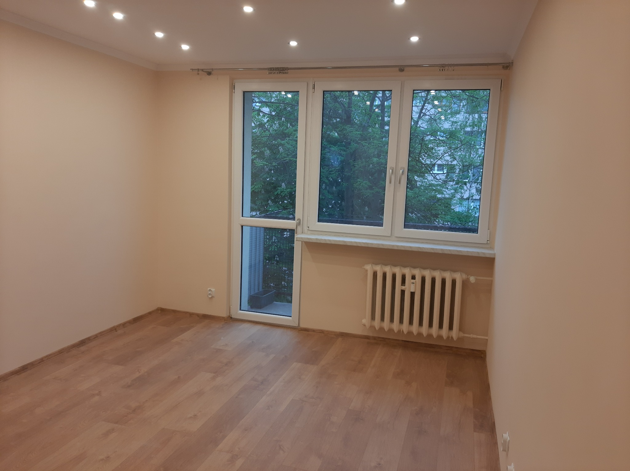 Pusty pokój po remoncie z jasnymi panelami podłogowymi, świeżo pomalowanymi beżowymi ścianami, białym grzejnikiem i oknem balkonowym z widokiem na zieleń.