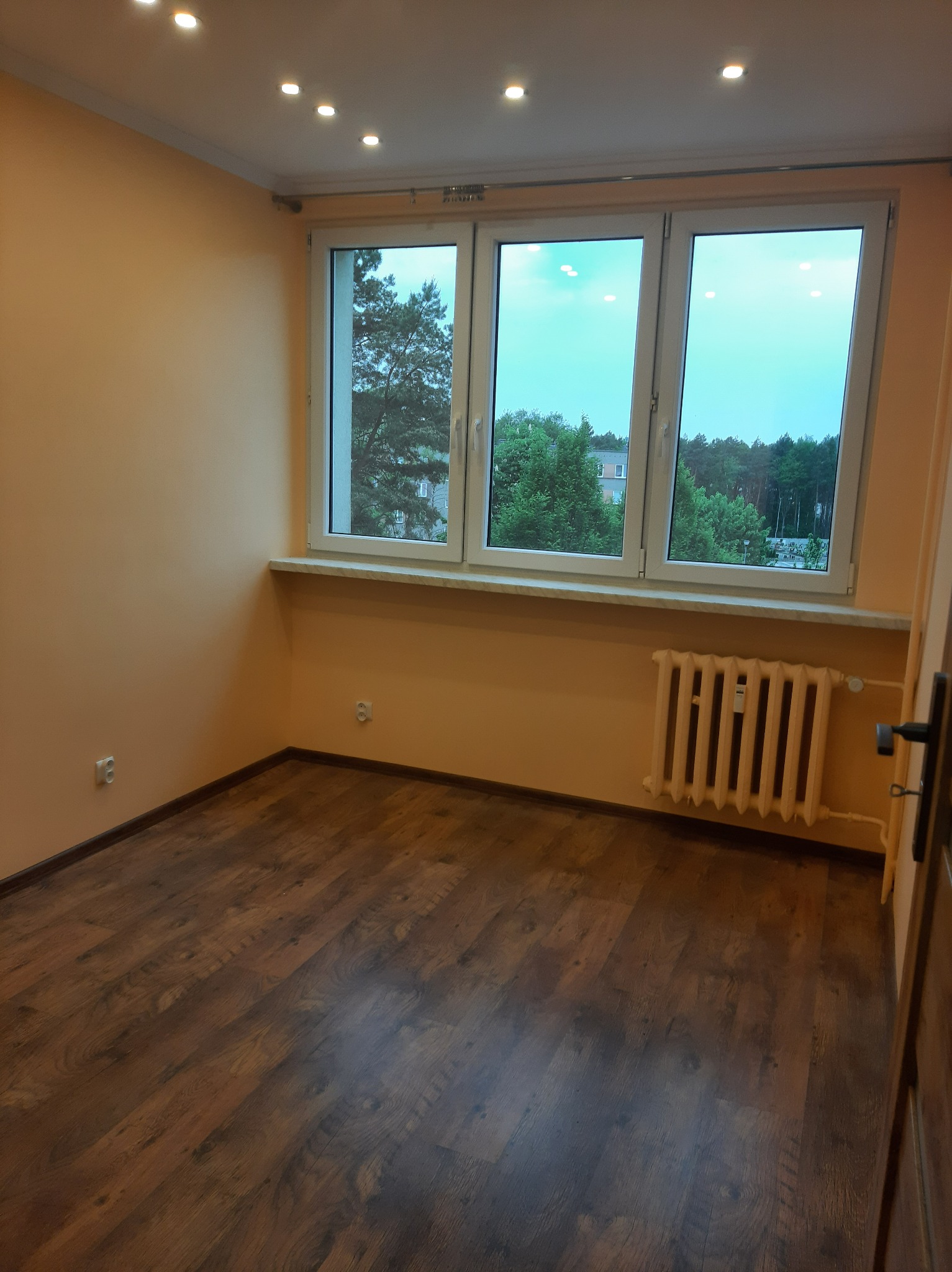 Pusty pokój z drewnianą podłogą, pomarańczowymi ścianami i białym grzejnikiem pod oknem z widokiem na zieleń.