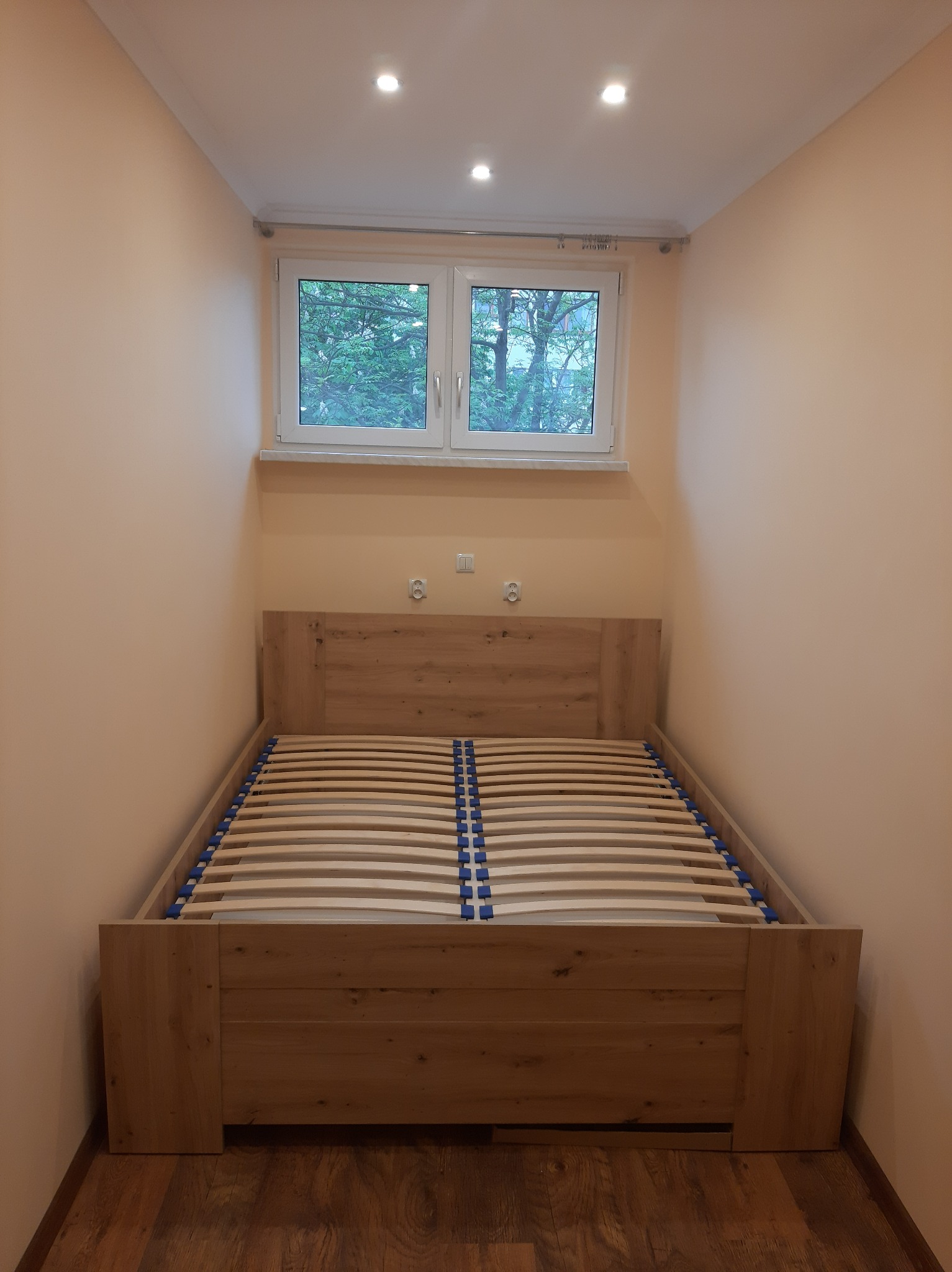 Wnętrze pokoju z łóżkiem z drewnianą ramą i listwami, świeżo pomalowane ściany w odcieniach beżu, widok z okna na zieleń drzew.