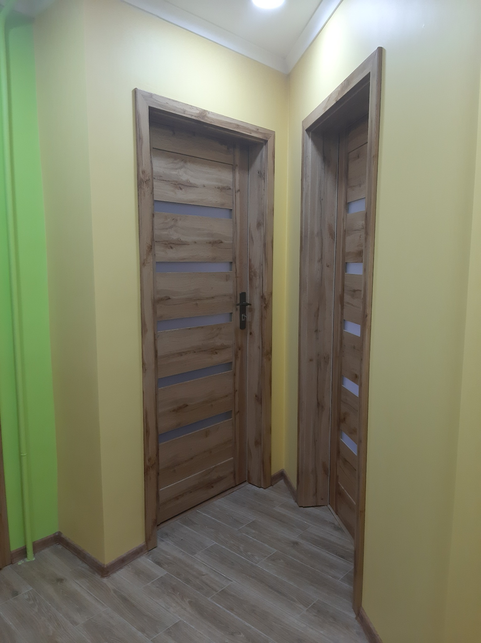 Dwa drewniane drzwi wewnętrzne z poziomymi przeszkleniami, zamontowane w żółtej ścianie, z widoczną podłogą z paneli imitujących drewno.