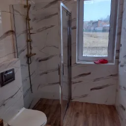 Nowoczesna łazienka z kabiną prysznicową typu walk-in, złotą armaturą, białymi płytkami imitującymi marmur z brązowymi żyłami, drewnopodobną podłogą, wiszącą miską WC i oknem z widokiem na domy...