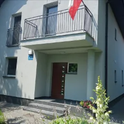 Jasnoszary dom jednorodzinny z balkonem, polską flagą, ciemnobrązowymi drzwiami wejściowymi i ozdobnym nasadzeniem roślin