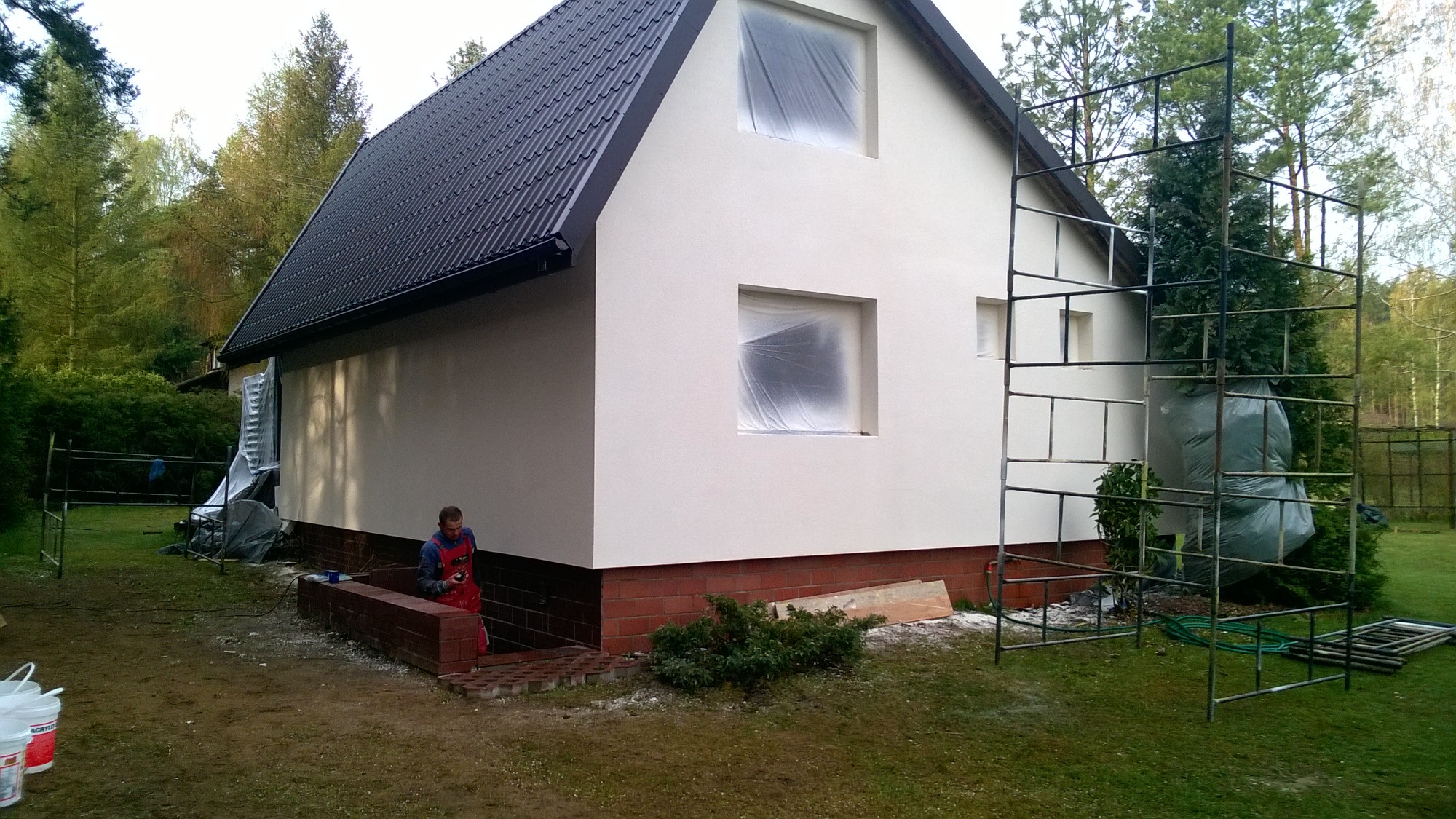 Dom jednorodzinny z nową, jasną elewacją; okna zabezpieczone folią malarską, widoczne rusztowanie i pracownik w czerwonym kombinezonie przygotowujący się do dalszych prac.