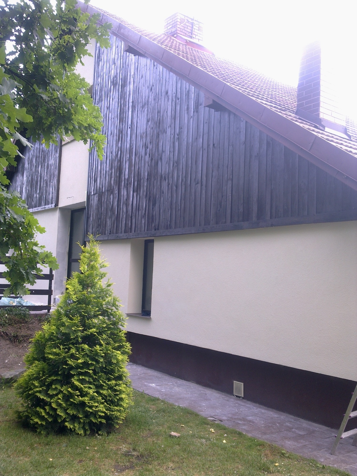 Elewacja domu z drewnianą okładziną i świeżo pomalowaną, jasną ścianą, widoczny fragment dachu i mała choinka na trawniku.