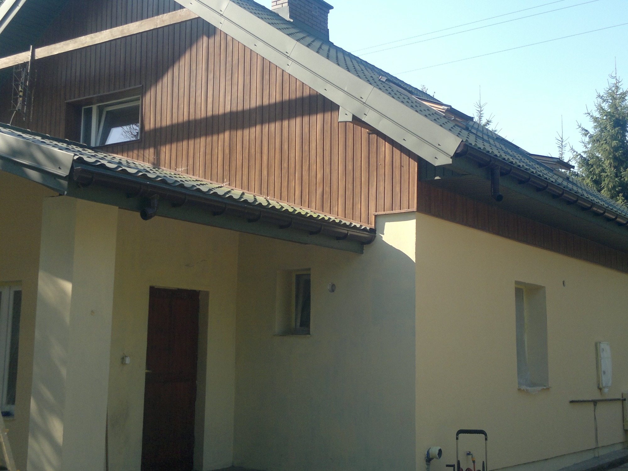 Dom jednorodzinny z elewacją częściowo wykończoną drewnianą boazerią, widoczna kremowa ściana z oknami i brązowe drzwi wejściowe, ciemna dachówka i zielone drzewa w tle.