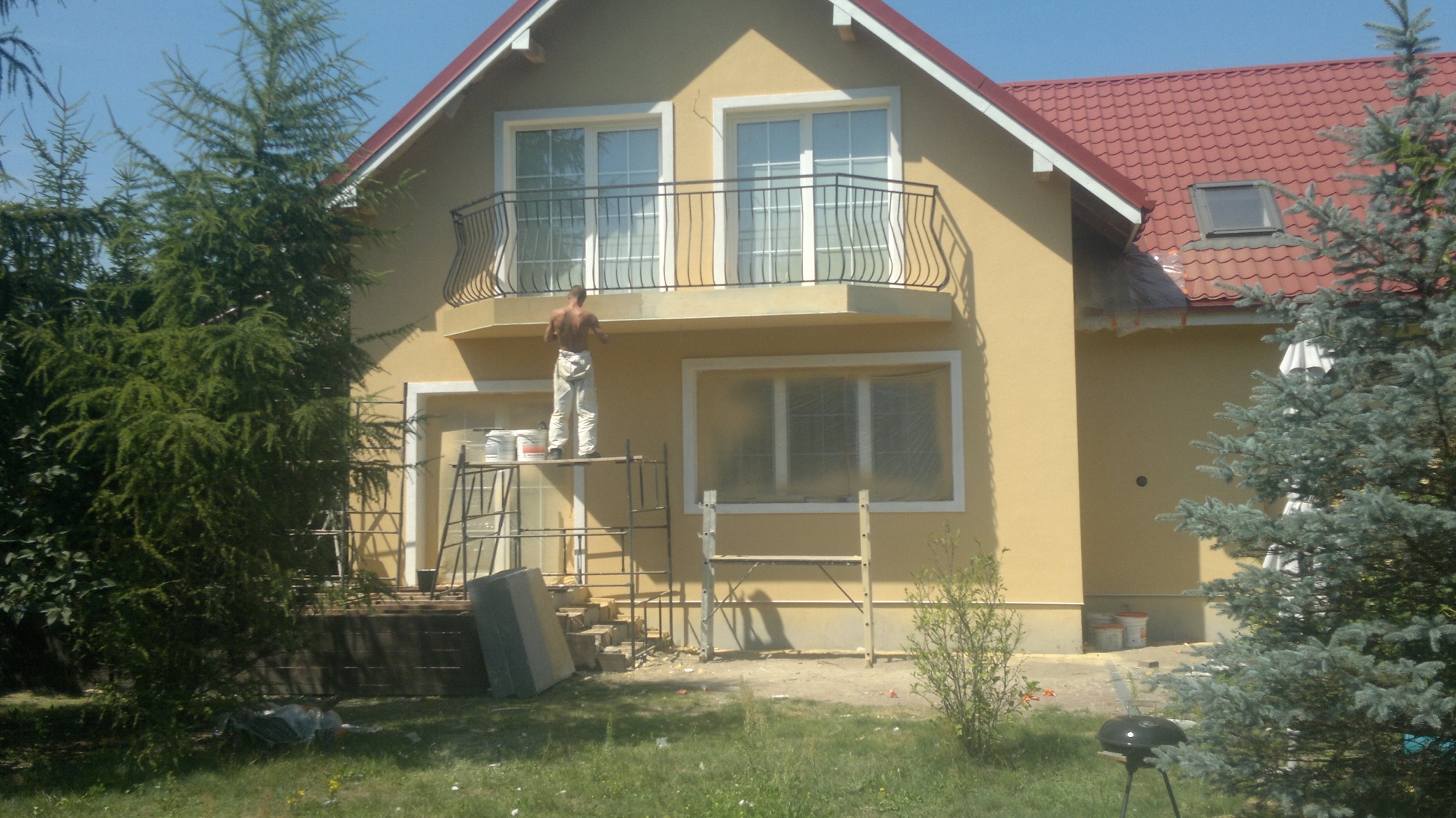 Mężczyzna bez koszulki, w spodniach roboczych, stoi na rusztowaniu przed domem z balkonem i maluje elewację. Okna zabezpieczone folią, widoczne puszki z farbą. Dom z czerwoną dachówką, otoczony...