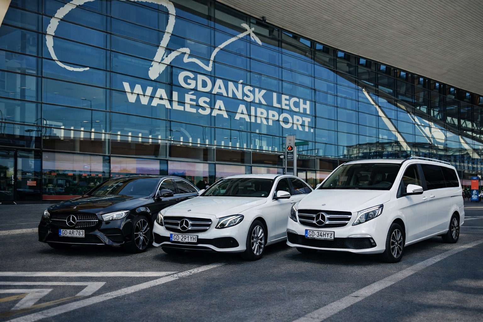 Flota trzech Mercedesów (czarny sedan, biała limuzyna i biały van) na tle lotniska w Gdańsku, prezentacja luksusowego transportu.