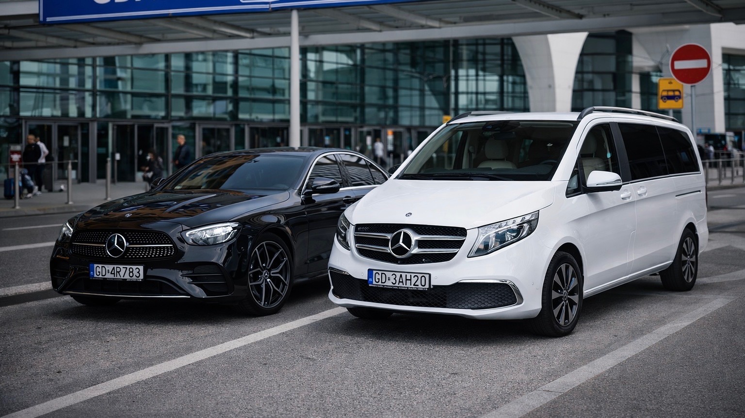Dwa luksusowe Mercedesy: czarna limuzyna i biały van, zaparkowane przed nowoczesnym budynkiem lotniska. Tablice rejestracyjne z Gdańska.
