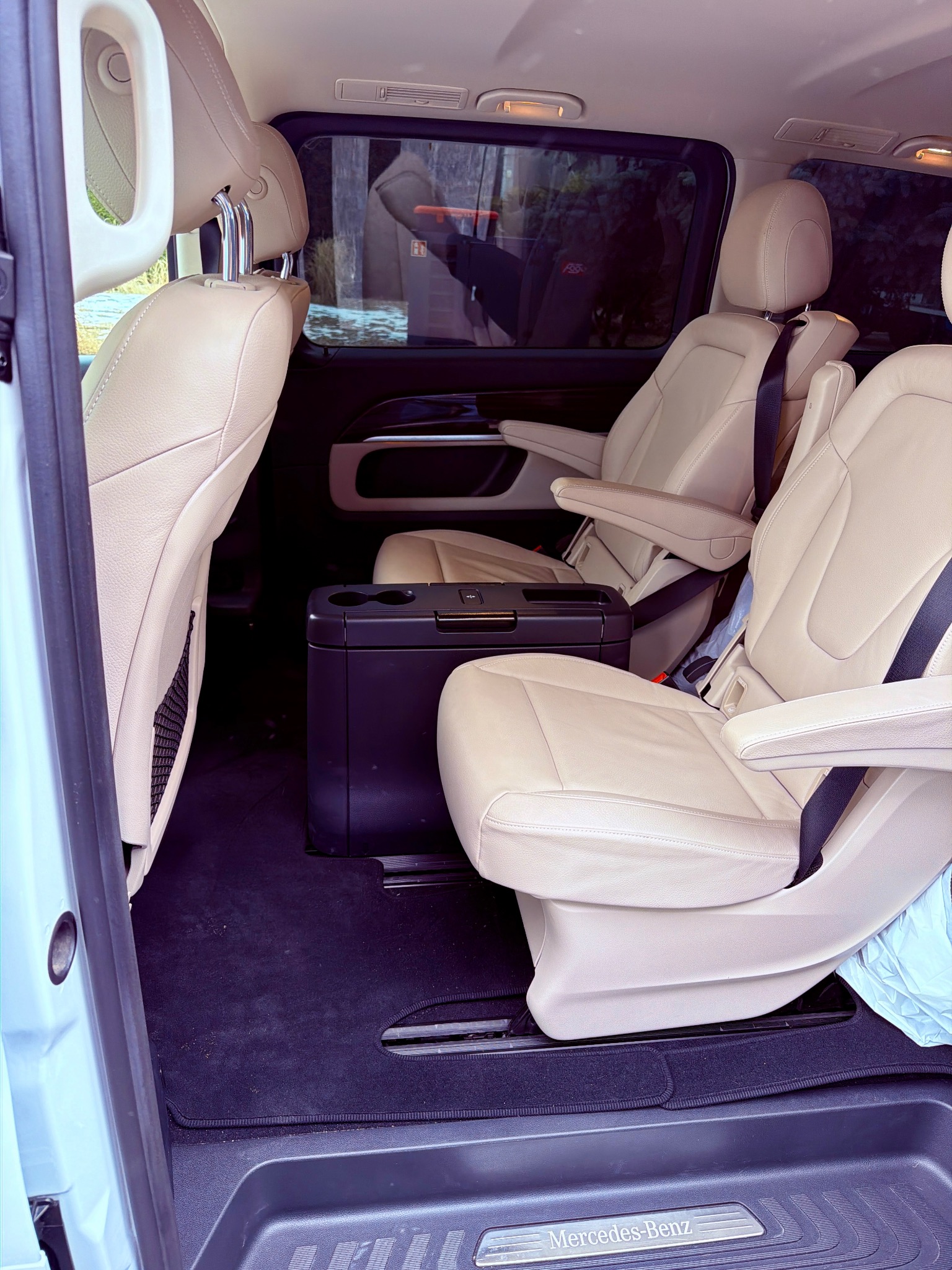 Wnętrze luksusowego busa Mercedes-Benz z beżowymi skórzanymi fotelami, czarną podłogą i lodówką między siedzeniami. Widok z otwartych drzwi.