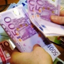 Dłonie trzymające plik banknotów 500 euro, obok klawiatura komputera i paczka banknotów 10 euro.