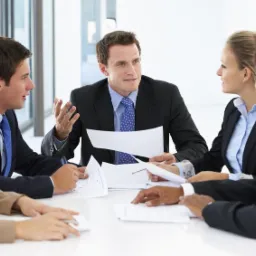 Zespół doradców w trakcie spotkania przy białym stole, analizujący dokumenty i omawiający strategię finansową w jasnym biurze.