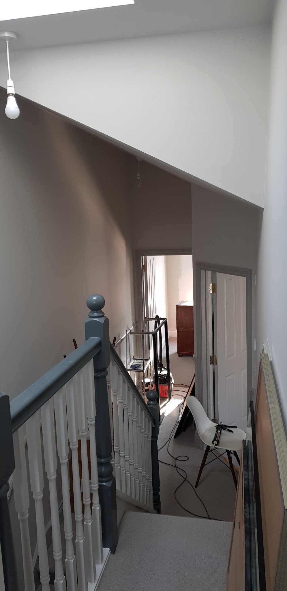 Klatka schodowa w trakcie malowania, widoczne pomalowane ściany w odcieniach szarości, białe schody, zabezpieczona folią podłoga, narzędzia malarskie na krześle i drabina malarska w tle.