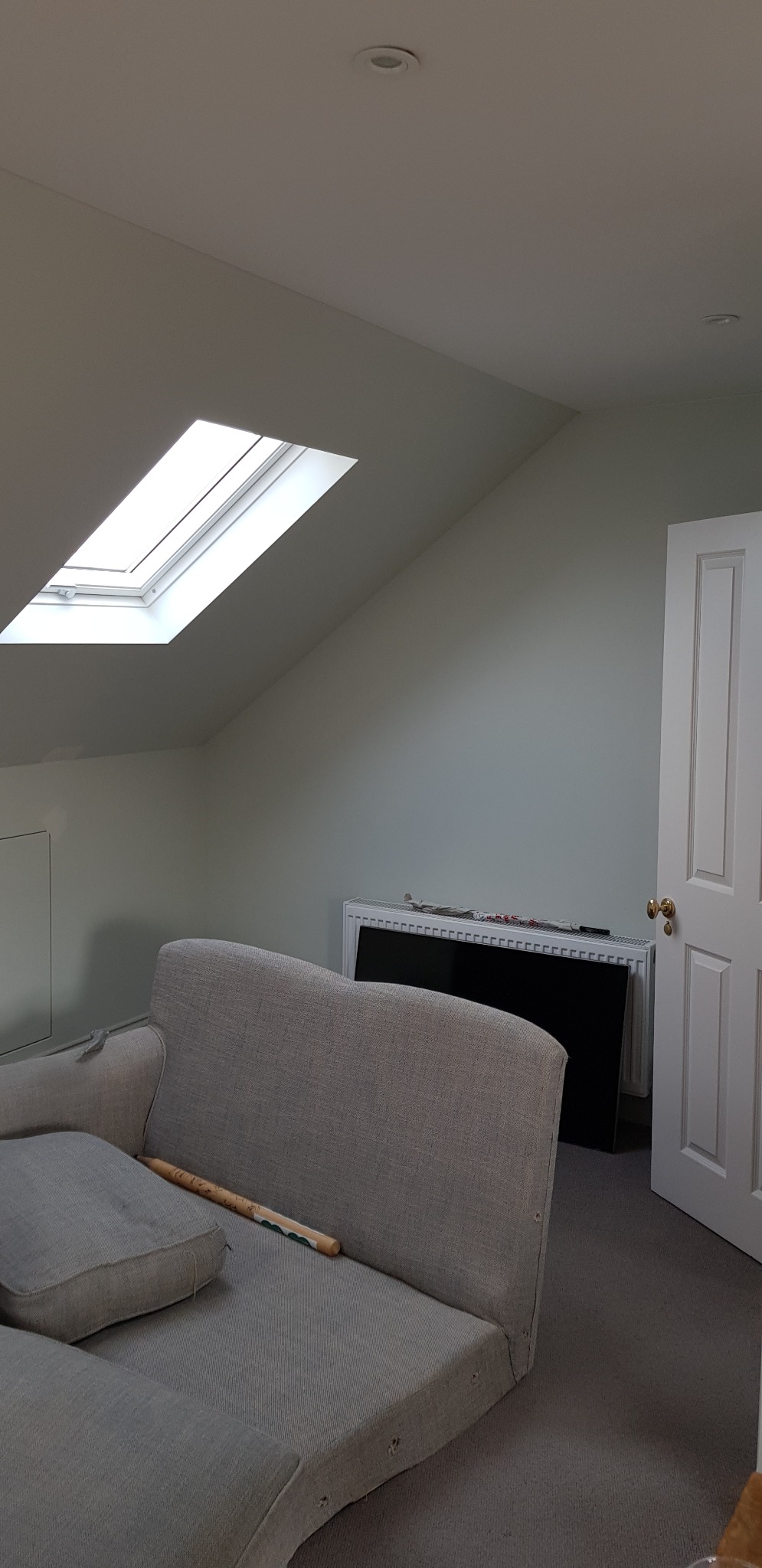Pokój na poddaszu z oknem dachowym i szarą sofą w trakcie remontu, ściany pomalowane na jasny kolor, widoczne przygotowania do prac wykończeniowych.