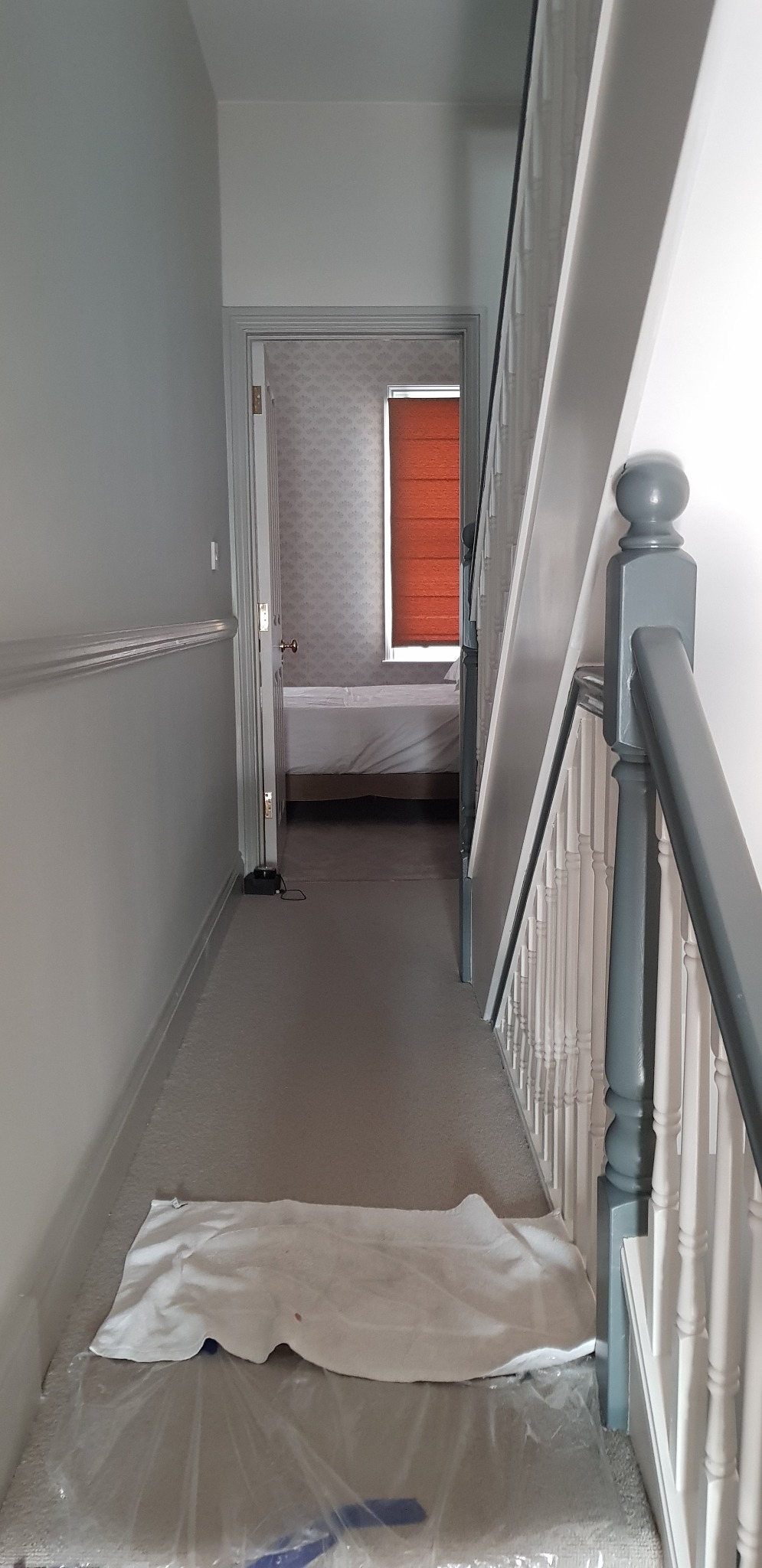 Korytarz w trakcie zabezpieczania przed pracami wykończeniowymi, widoczna folia ochronna na podłodze i ręcznik przy schodach, w tle otwarte drzwi do pokoju z roletą w oknie.