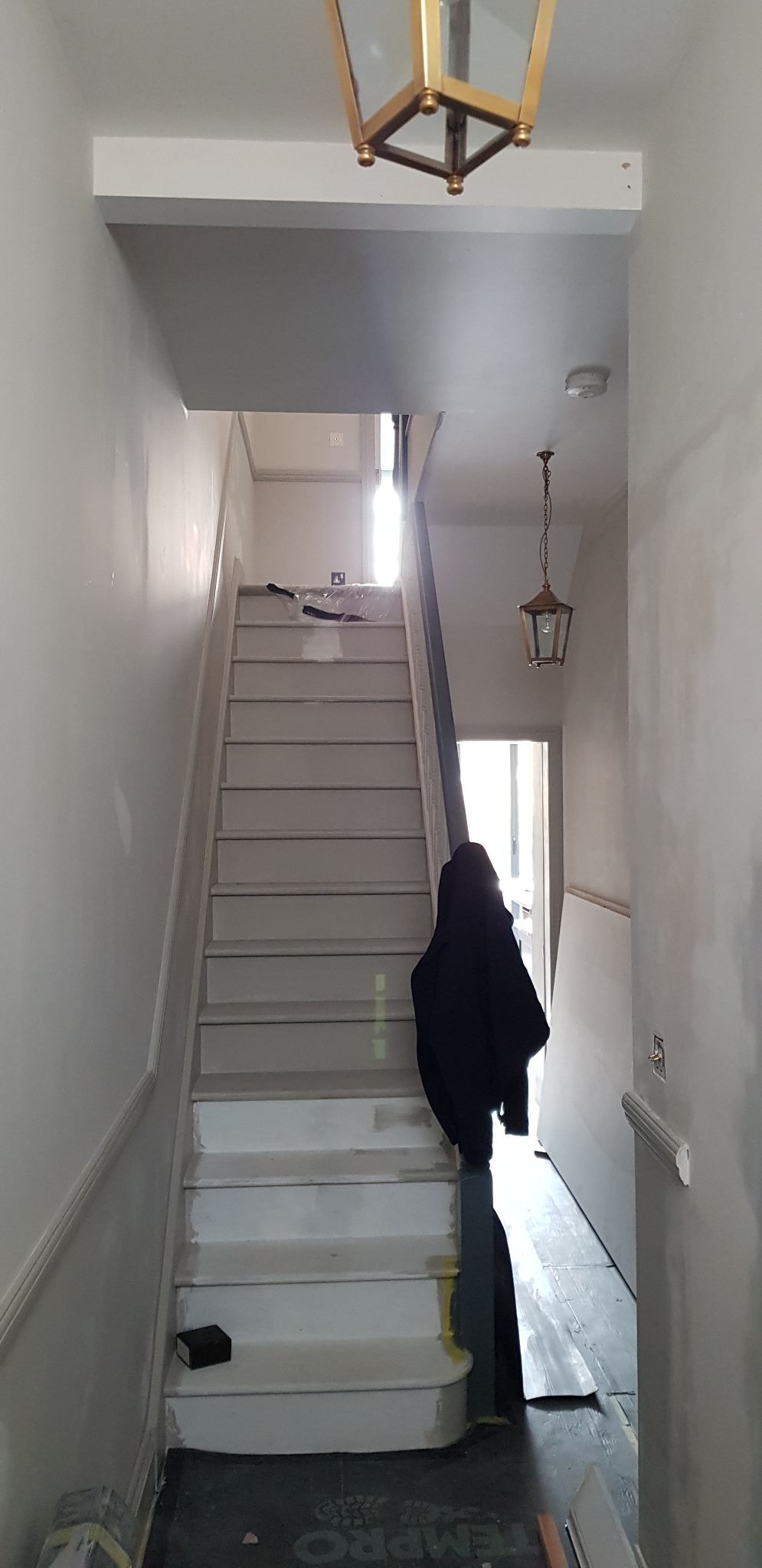 Klatka schodowa w trakcie prac wykończeniowych: świeżo pomalowane schody w odcieniach szarości i bieli, widoczne ślady po taśmie malarskiej, wisząca lampa w stylu retro i czarny płaszcz na poręczy.