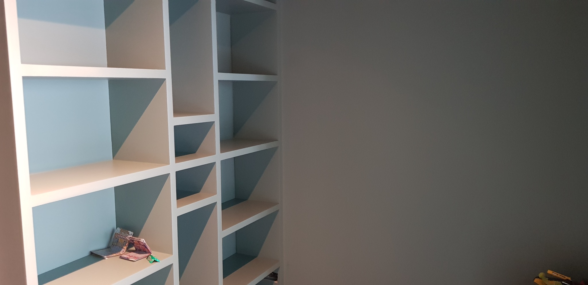 Biały regał z półkami, pomalowany na kolor błękitno-beżowy, z kilkoma przedmiotami na jednej z półek, ustawiony przy szarej ścianie.