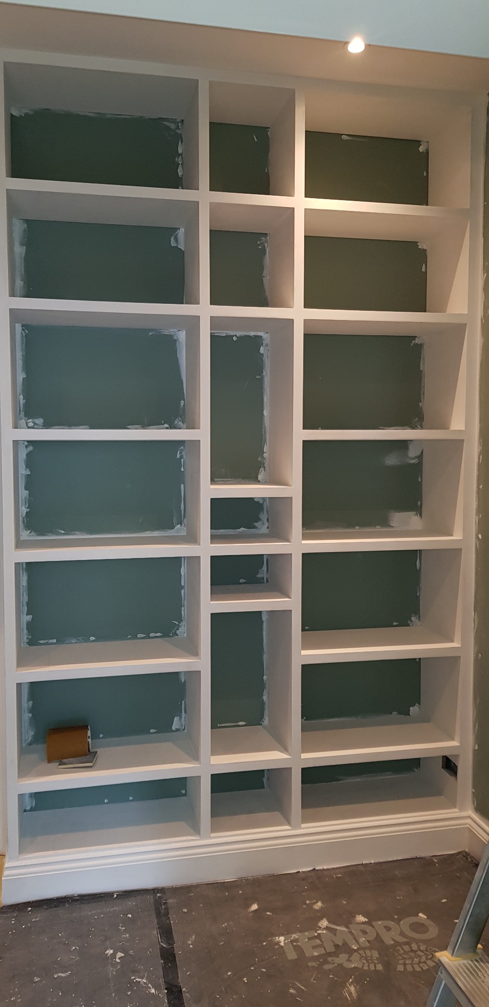 Biały regał wnękowy z zielonym tłem półek, w trakcie prac wykończeniowych, widoczne ślady szpachli przy krawędziach, na podłodze folia ochronna.