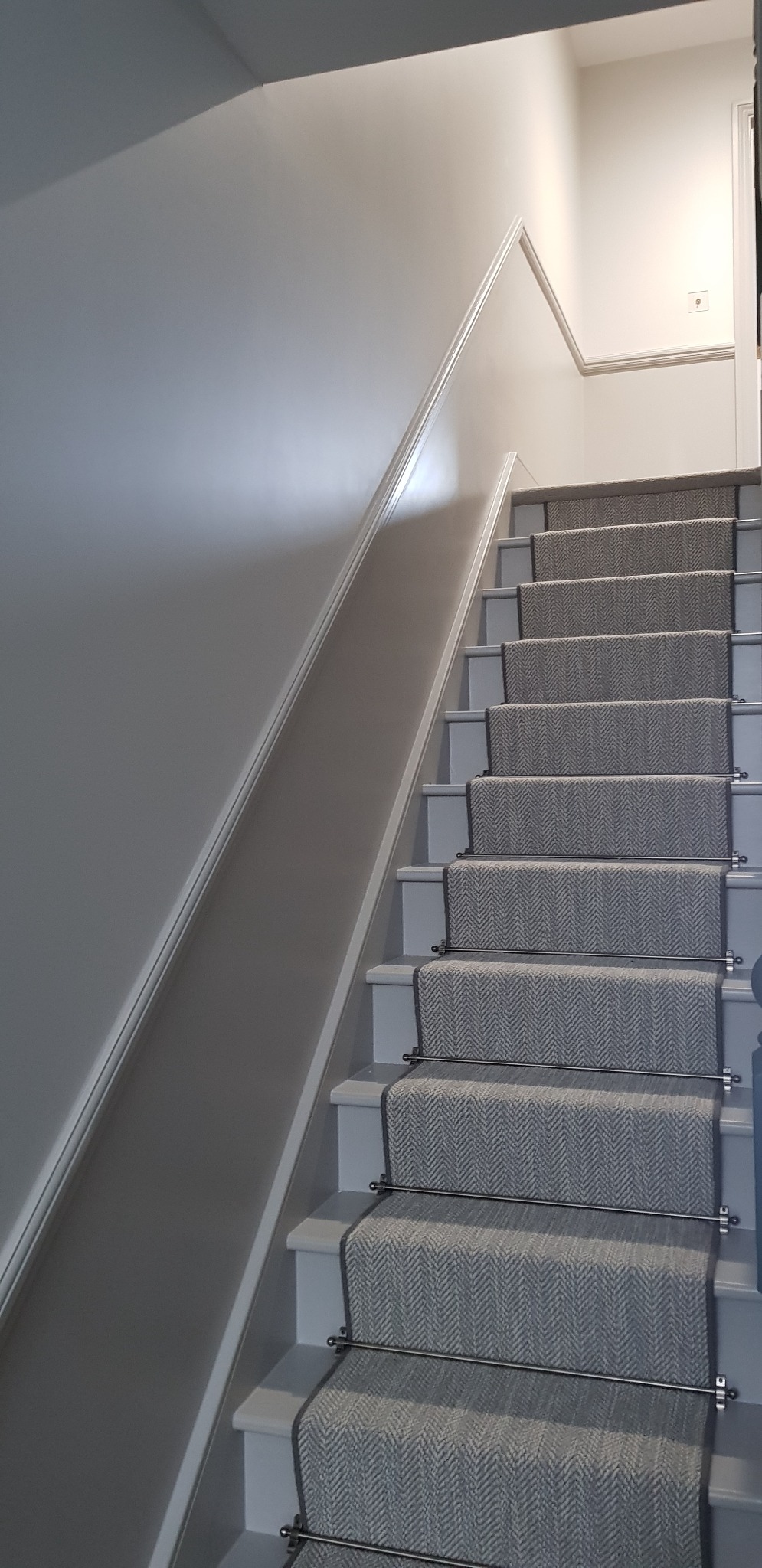 Schody w odcieniach szarości z dywanem w jodełkę, oświetlone światłem padającym z góry.
