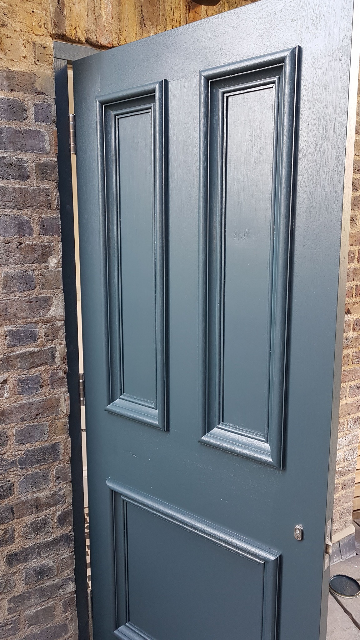 Pomalowane na szaro-zielony kolor, częściowo otwarte drzwi z dekoracyjnymi panelami, widoczne zawiasy i fragment muru z cegły.