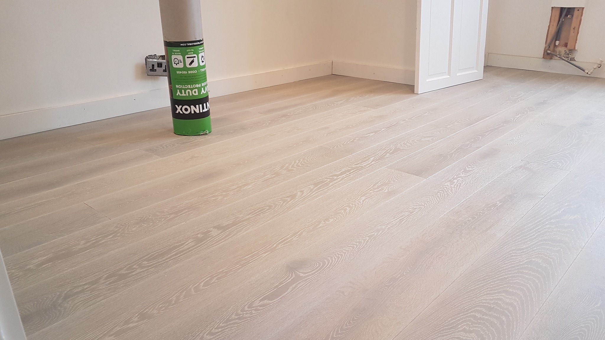 Świeżo położona, jasna, drewniana podłoga z widoczną rolką ochronnej folii podłogowej przy ścianie i niedokończonym fragmentem ściany z instalacją.