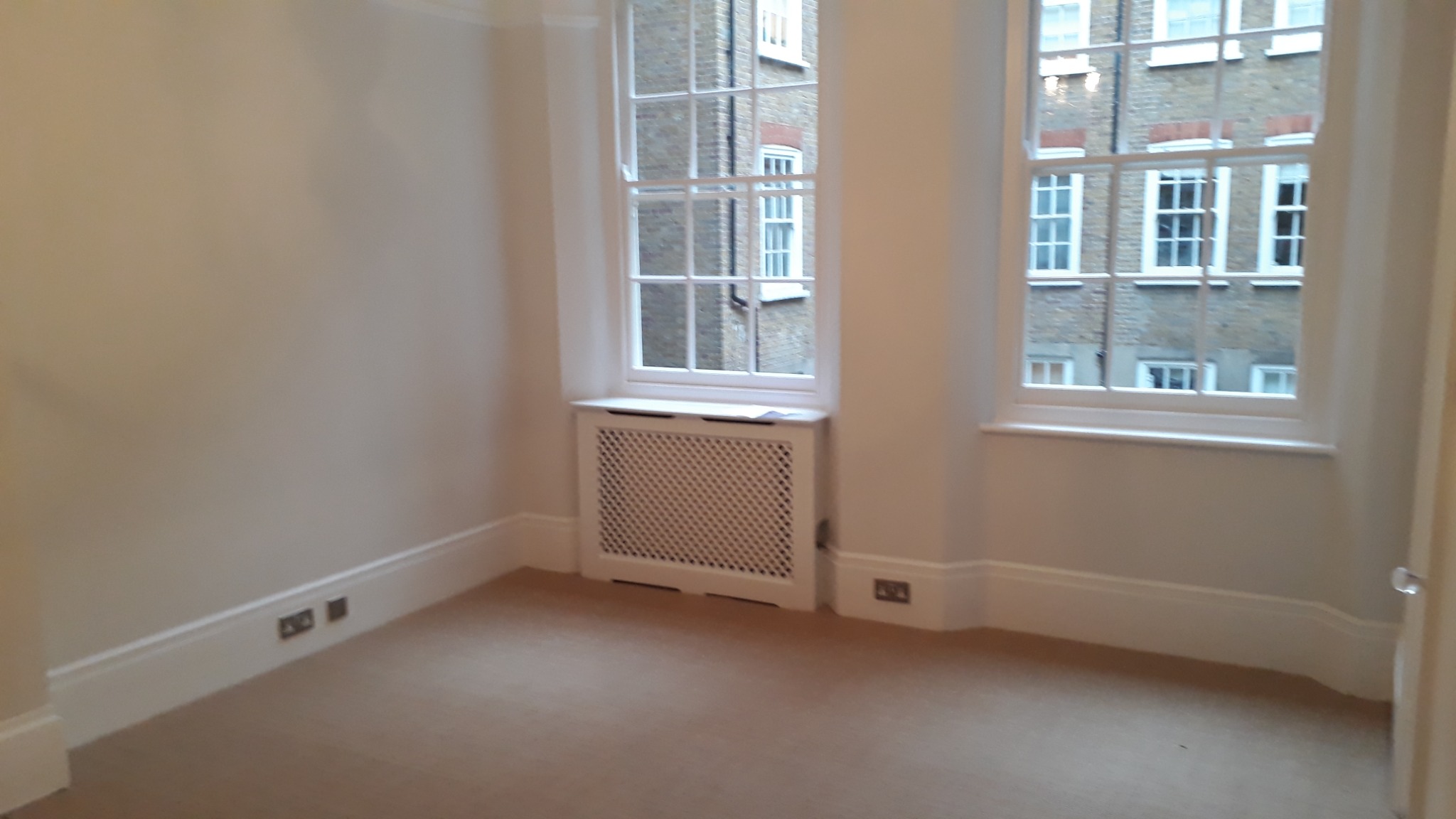 Pusty pokój po malowaniu z dwoma oknami z widokiem na budynek z cegły, grzejnik zakryty białą osłoną, beżowa wykładzina, białe listwy przypodłogowe i ściany w odcieniach beżu.