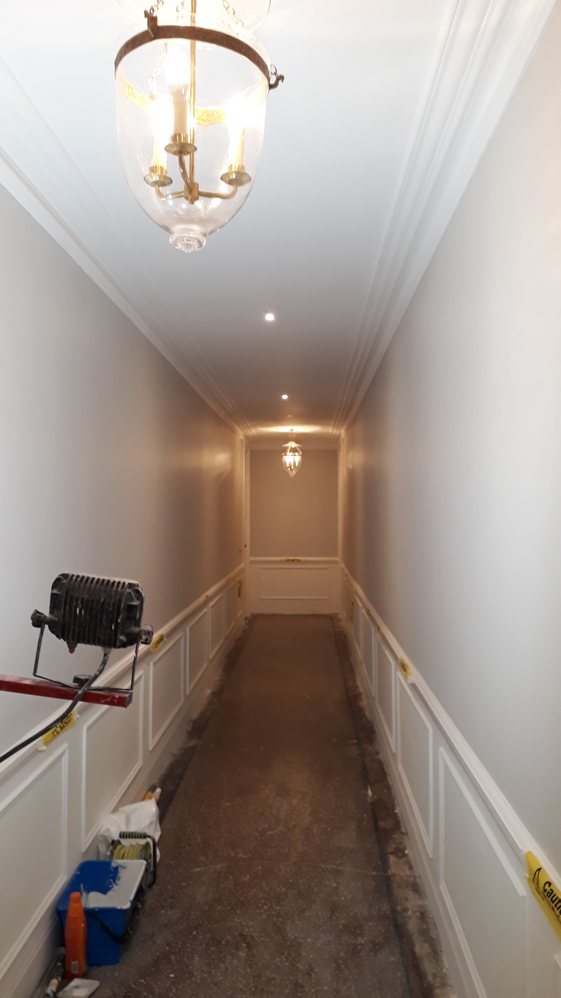Korytarz w trakcie odświeżania: świeżo pomalowane ściany w jasnym odcieniu, zabezpieczone taśmą, widoczny reflektor budowlany i akcesoria malarskie na podłodze.