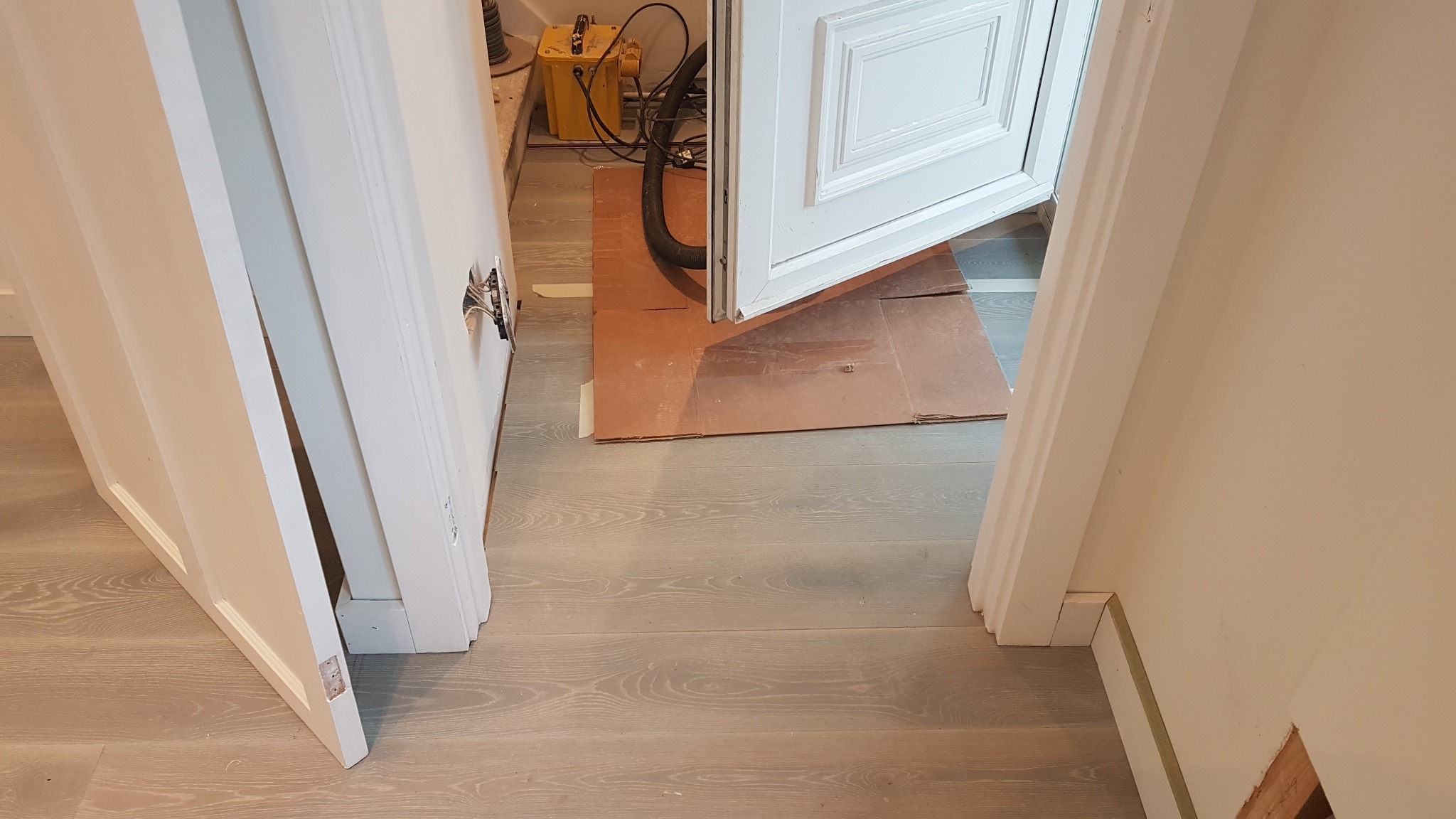 Otwarta biała stolarka drzwiowa z widocznym zabezpieczeniem podłogi kartonem, w tle żółty transformator i czarny przewód