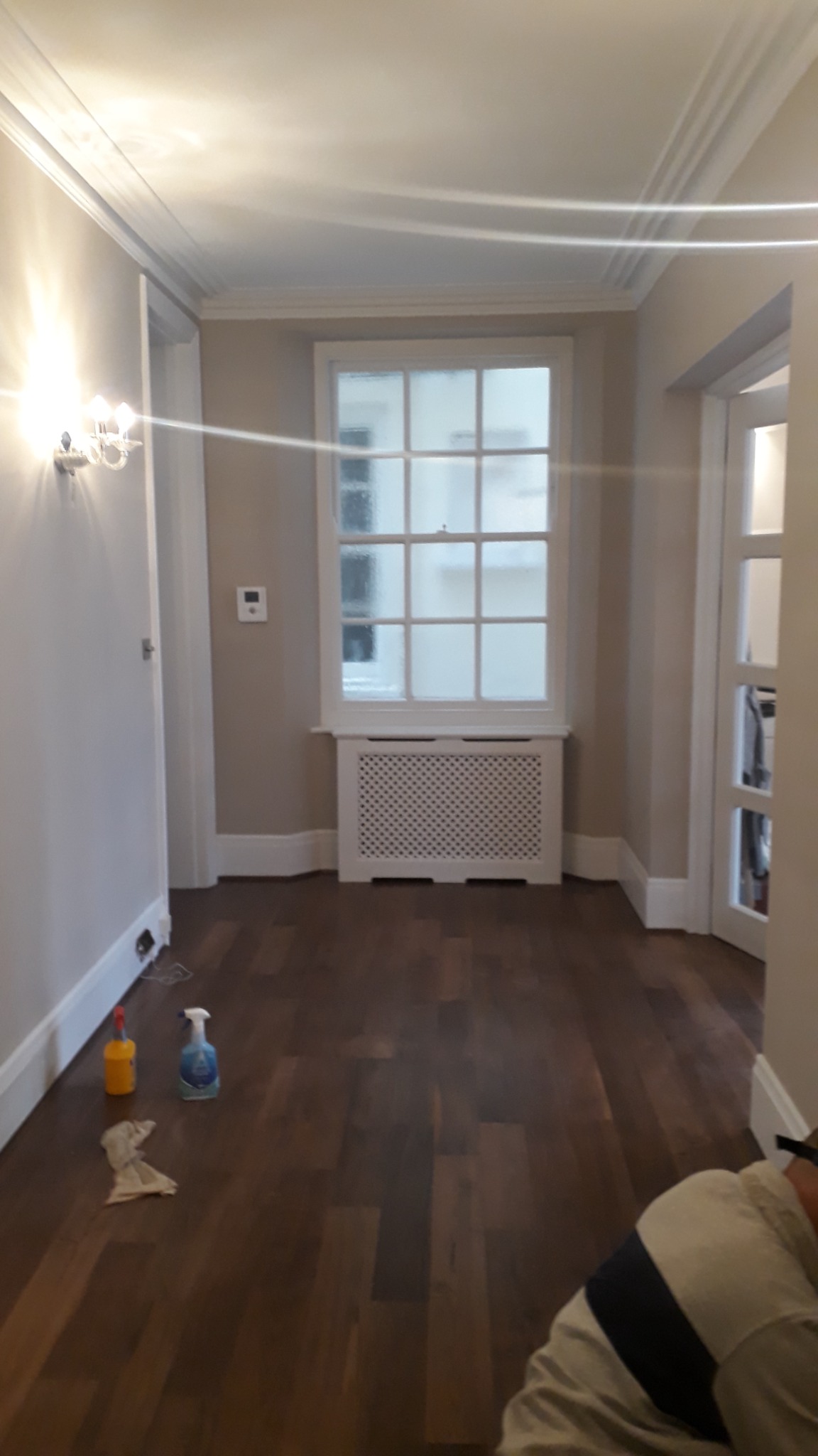 Wnętrze pokoju po odświeżeniu: świeżo pomalowane ściany w odcieniach beżu, ciemna drewniana podłoga, biała stolarka okienna i listwy przypodłogowe, widoczne butelki ze środkami czyszczącymi.