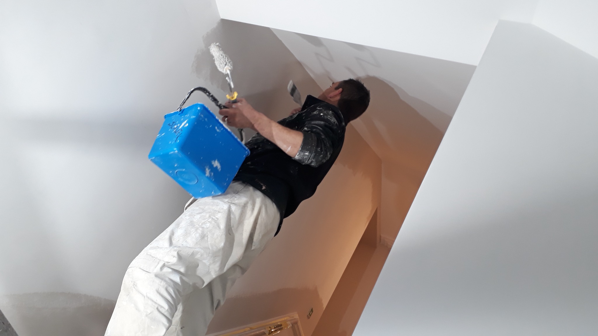 Mężczyzna w ubraniu roboczym maluje białą ścianę na poddaszu za pomocą wałka i niebieskiego pojemnika z farbą.