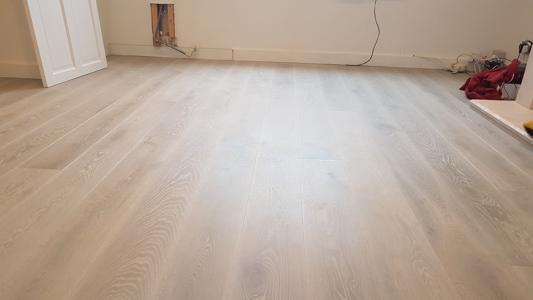 Jasna, drewniana podłoga po renowacji, widoczne ślady po niedawno zakończonych pracach, niezabezpieczone kable i otwór w ścianie.