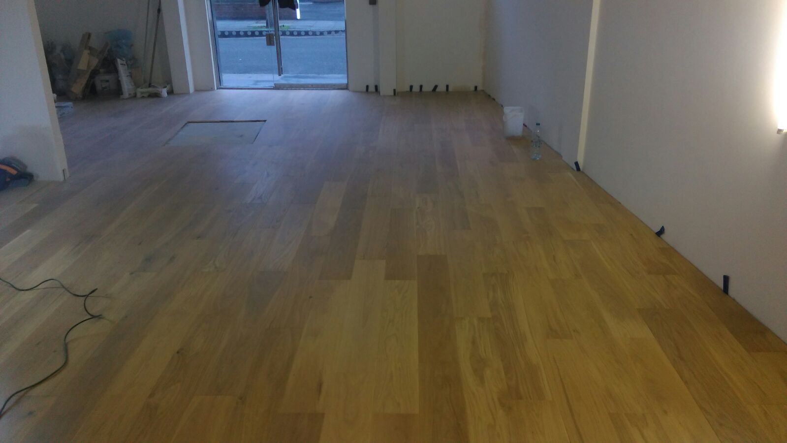 Jasna, drewniana podłoga ułożona w nowym biurze, z widocznymi dystansami przy ścianie i otwartym otworem w podłodze, sugerującym trwające prace wykończeniowe.