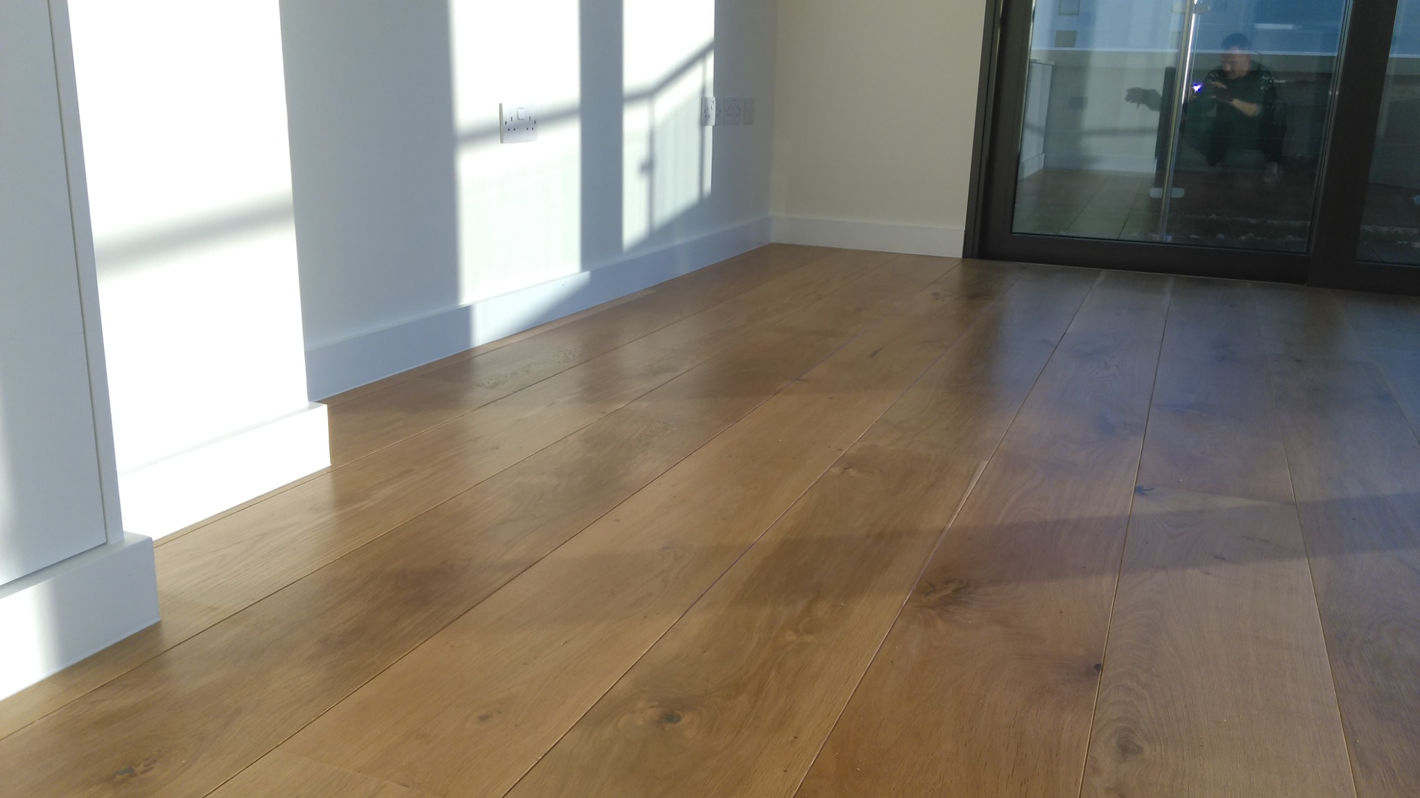 Ułożona drewniana podłoga z widocznymi słojami, biała listwa przypodłogowa, cień na ścianie i odbicie osoby w szybie balkonu.