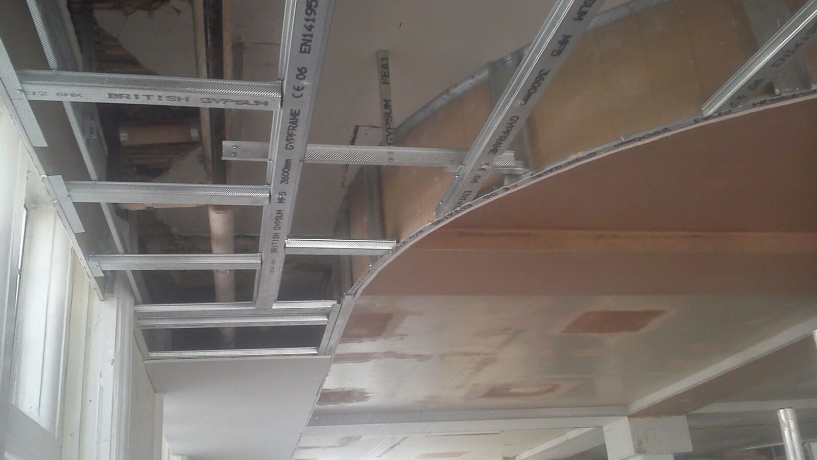 Sufit podwieszany z metalowej konstrukcji i płyt gipsowo-kartonowych w trakcie montażu, widoczne profile British Gypsum i instalacje nad sufitem.