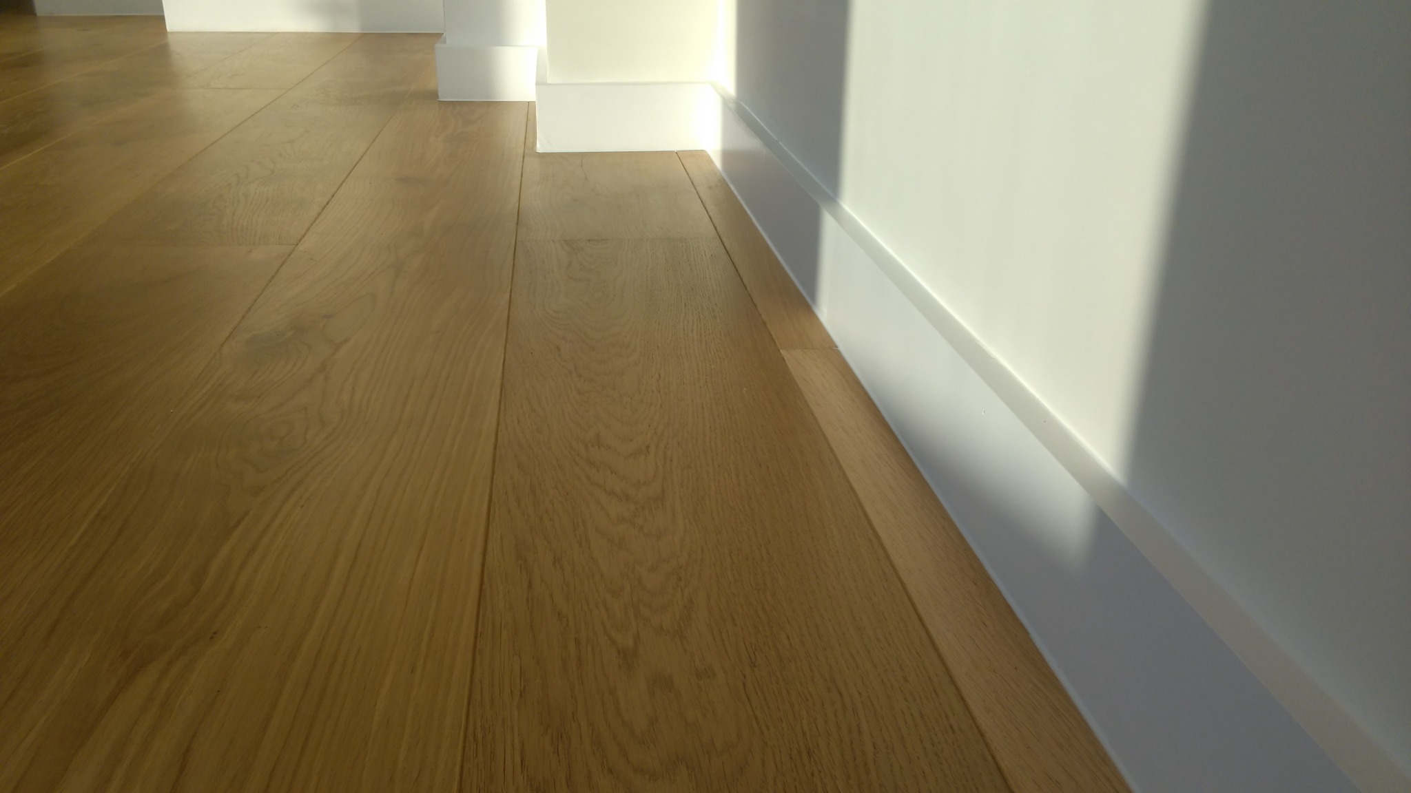 Jasny parkiet ułożony przy białej ścianie z przypodłogową listwą, oświetlony popołudniowym światłem.