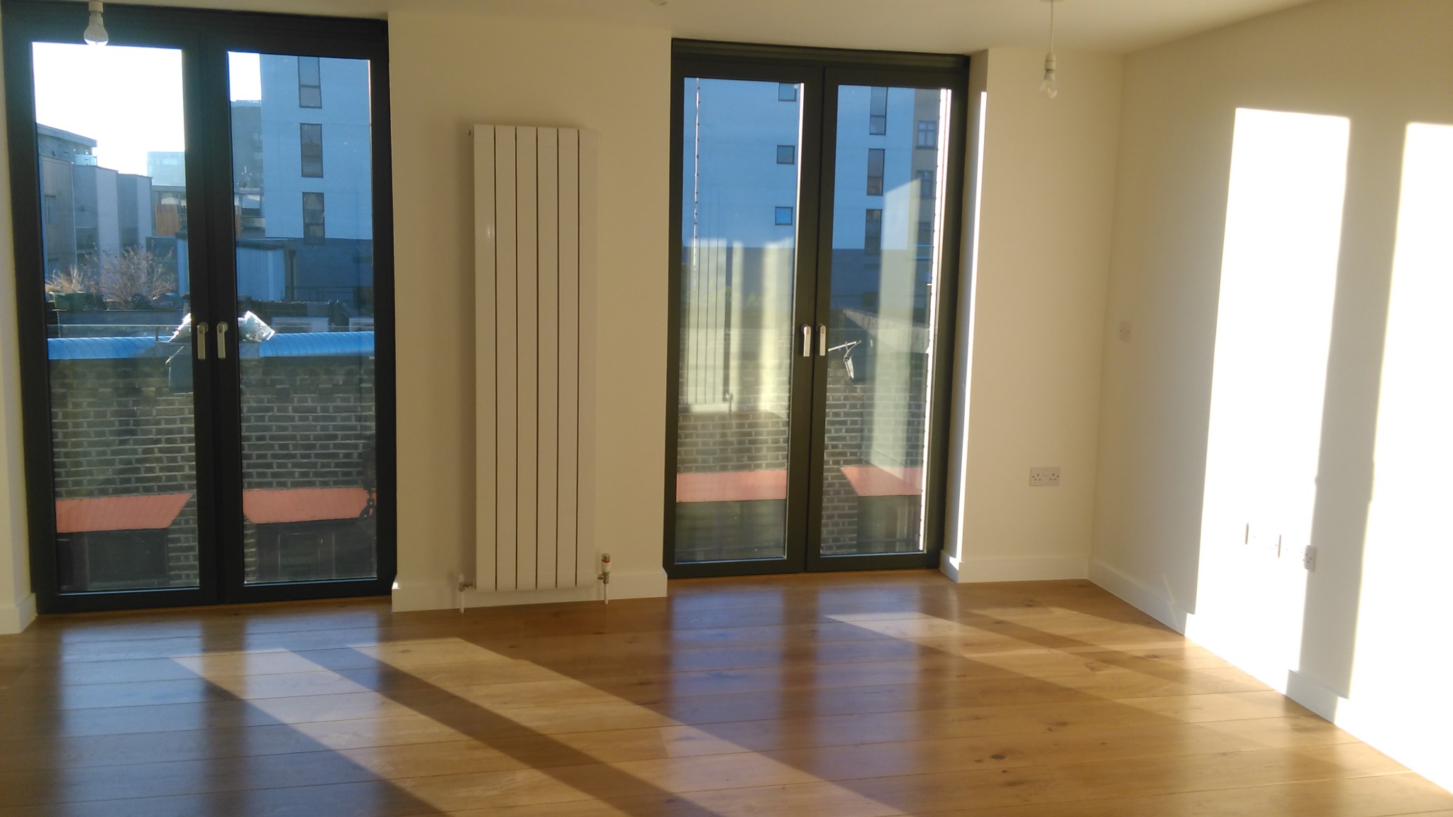 Jasne, puste pomieszczenie z drewnianą podłogą, dużymi oknami balkonowymi z czarnymi ramami i białym grzejnikiem ściennym, zalane popołudniowym słońcem.