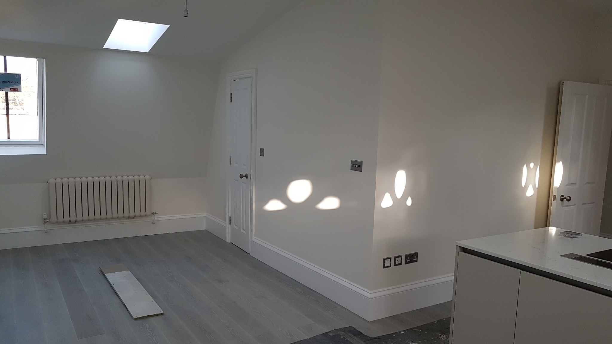 Jasne, nowocześnie wykończone wnętrze z szarymi panelami podłogowymi, białymi ścianami i grzejnikiem. Promienie słoneczne tworzą abstrakcyjne wzory na ścianie.