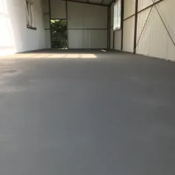 Świeżo wylana, gładka, szara posadzka w hali magazynowej z metalowymi ścianami, widoczne okno i otwarte drzwi na końcu pomieszczenia.