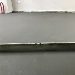 Świeżo wylana, szara posadzka wyrównywana długą poziomicą w pomieszczeniu z białymi ścianami i wystającymi rurami.