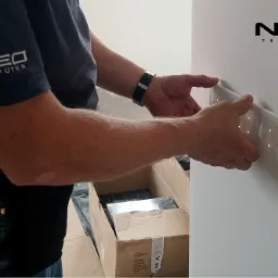 Instalator montuje panel sterowania inteligentnego domu z podświetlanymi przyciskami na białej ścianie, widoczny karton z elementami instalacji i logo firmy NEO na koszulce pracownika.