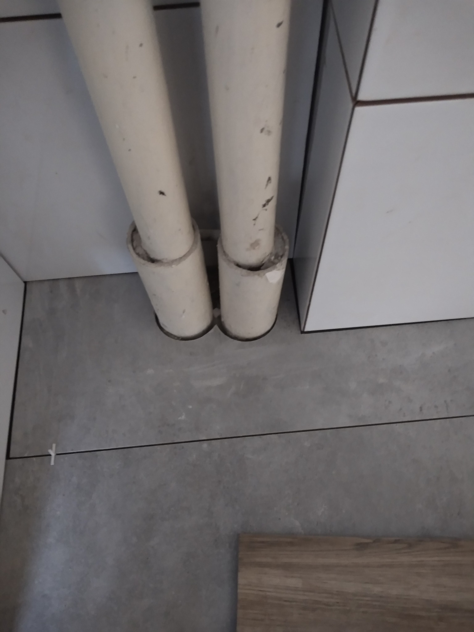 Szare płytki podłogowe ułożone wokół dwóch białych rur, z widocznym fragmentem białej szafki i krzyżykiem dystansowym w spoinie.