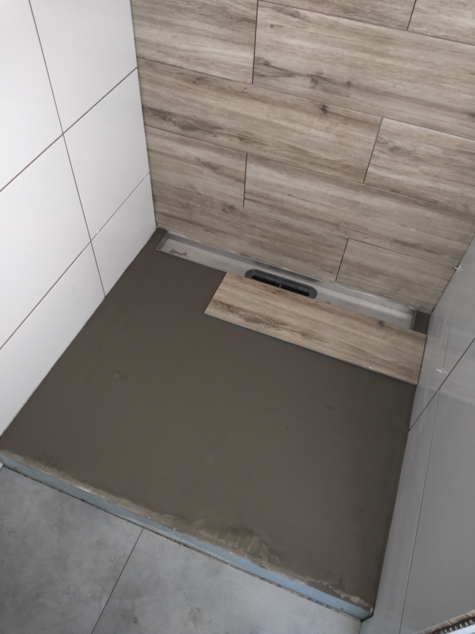 Przygotowanie podłoża pod prysznic z odpływem liniowym, z widocznymi płytkami imitującymi drewno na ścianie i białymi, kwadratowymi płytkami na sąsiedniej ścianie.