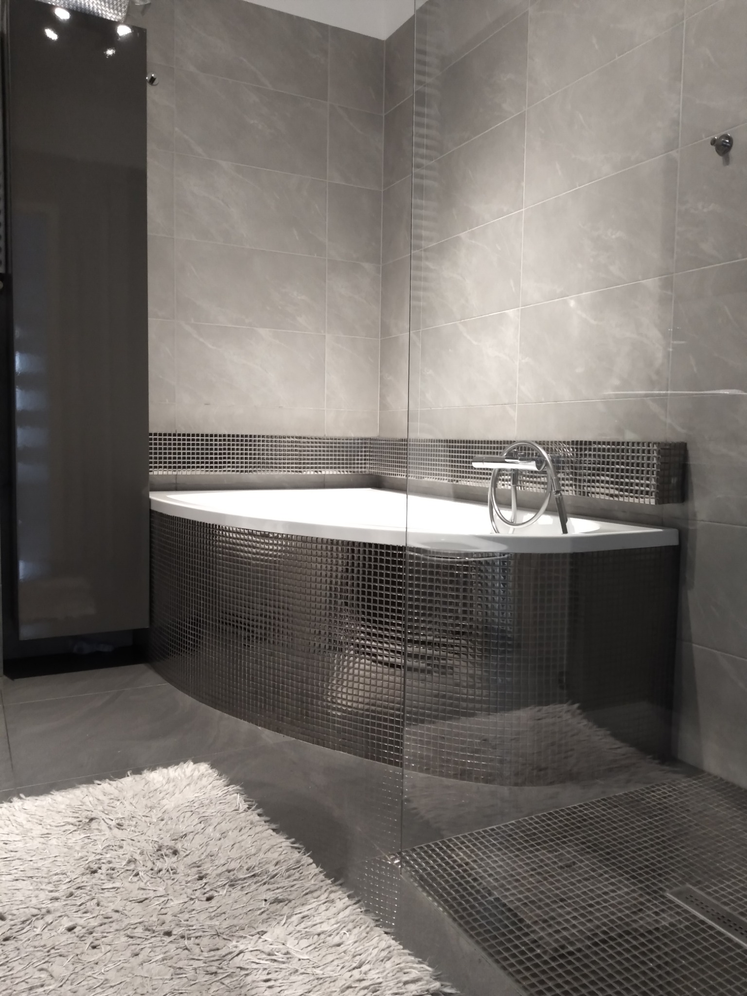 Nowoczesna łazienka w odcieniach szarości z wanną obudowaną mozaiką, szklaną kabiną prysznicową i puszystym dywanikiem.