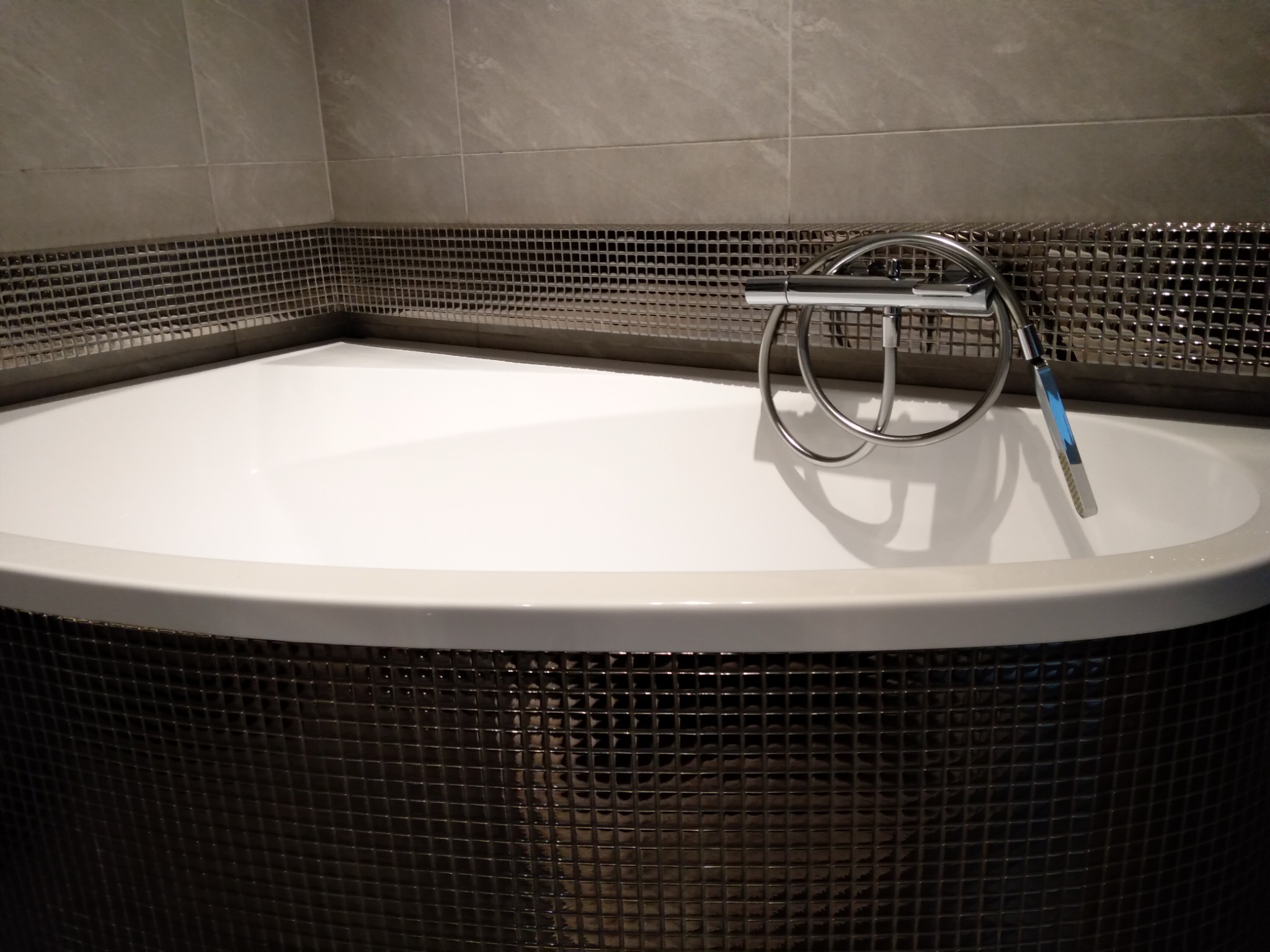 Biała, narożna wanna obłożona błyszczącą, ciemną mozaiką, z chromowaną baterią i słuchawką prysznicową na tle beżowych płytek.