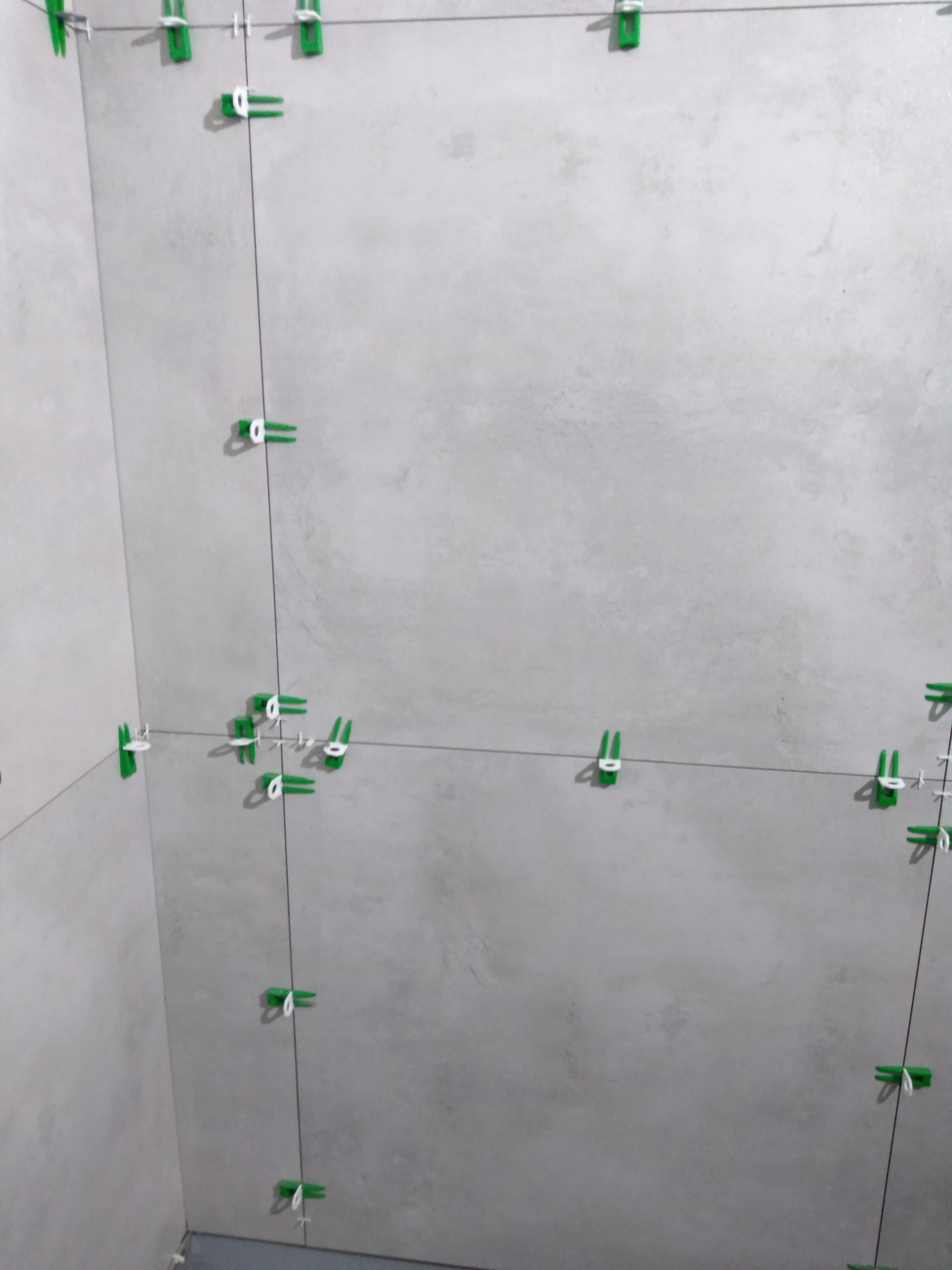 Świeżo ułożone szare płytki w rogu pomieszczenia, z użyciem zielonych klinów i sznurka do poziomowania, widoczne delikatne nierówności na powierzchni płytek.