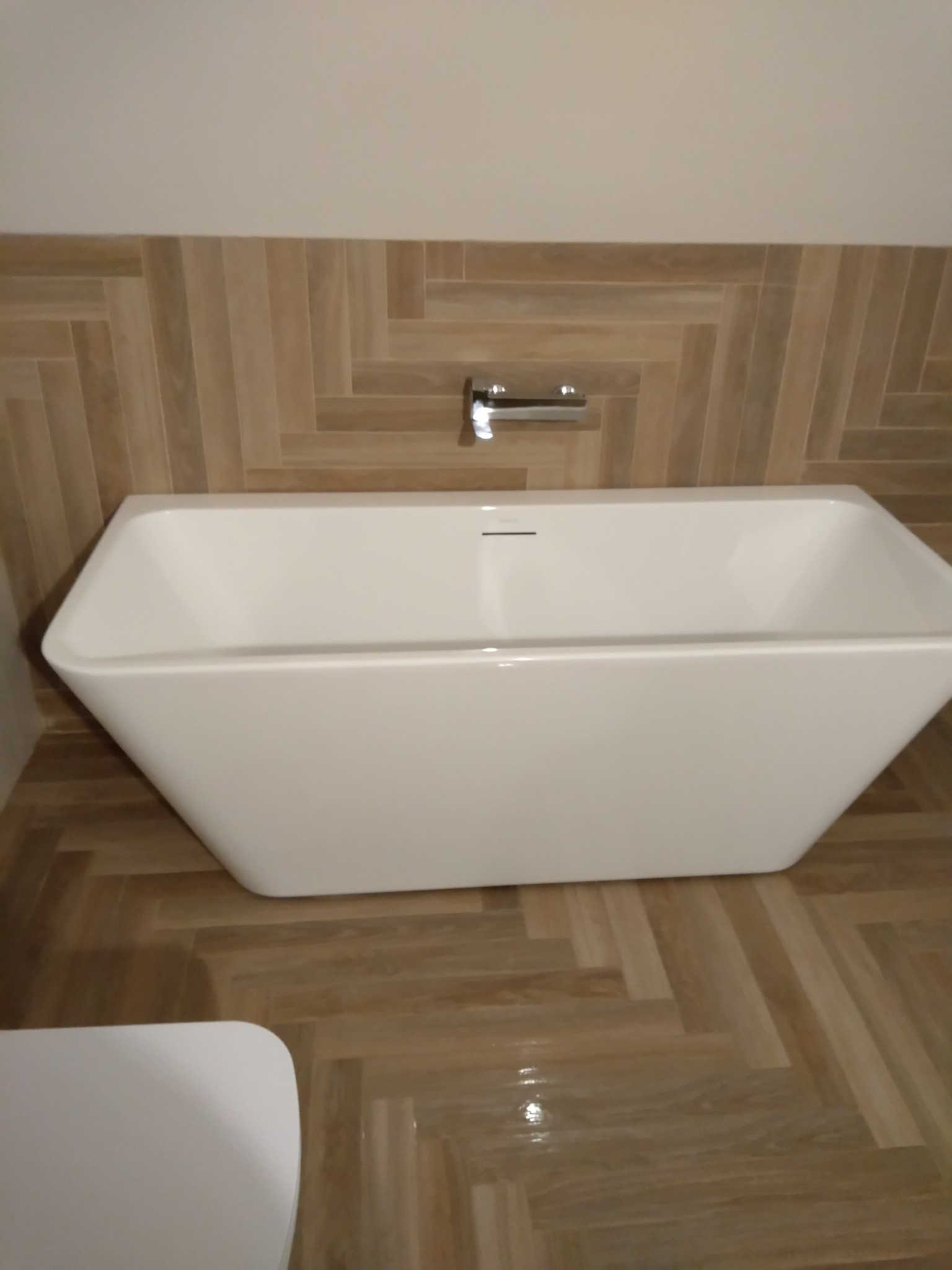 Nowoczesna, wolnostojąca wanna o geometrycznym kształcie, umieszczona na podłodze i ścianie wyłożonej płytkami imitującymi drewno w jodełkę, z minimalistyczną baterią ścienną.