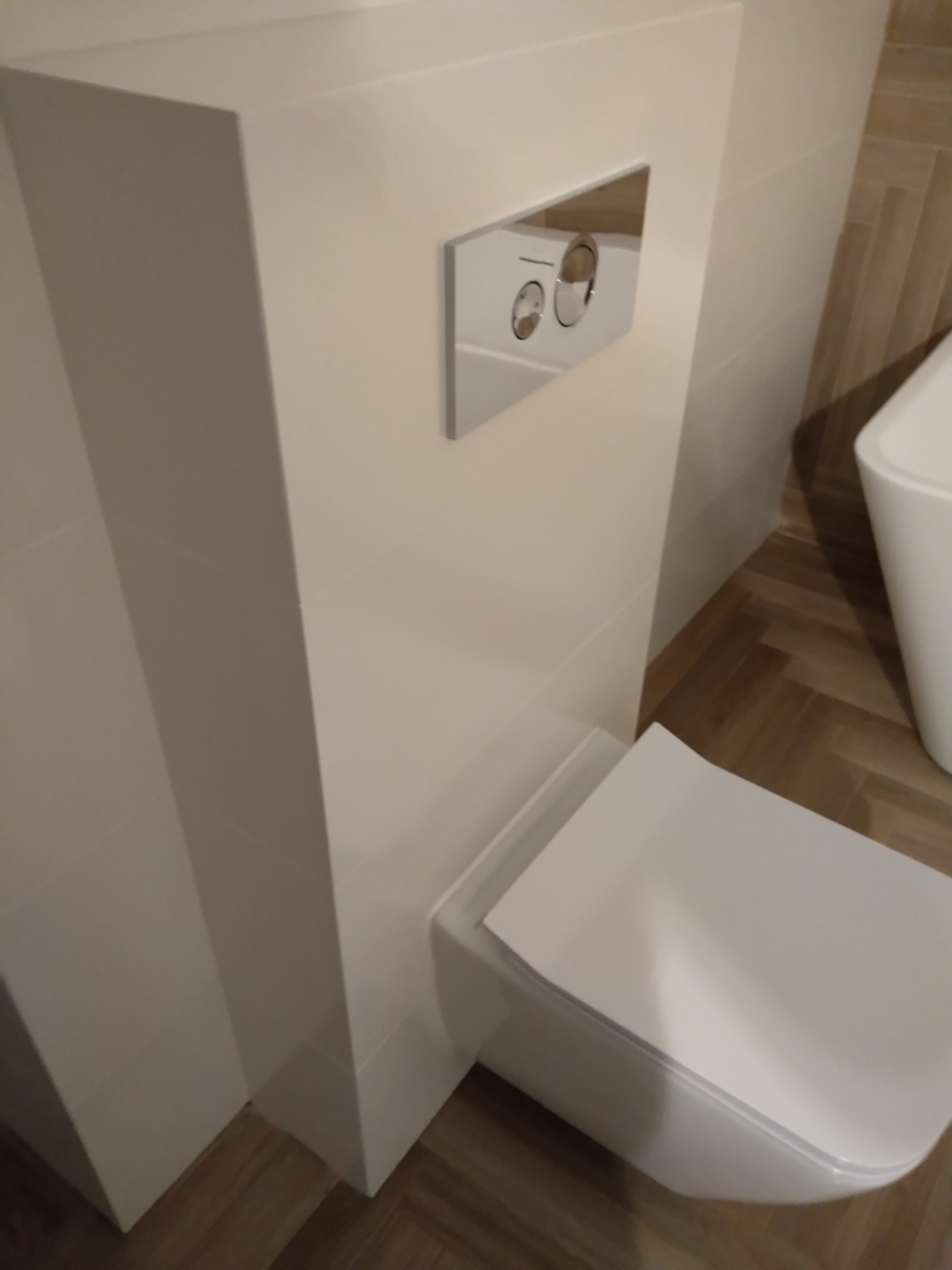 Podwieszana toaleta z zabudowaną spłuczką na tle płytek imitujących drewno, widoczne chromowane przyciski spłukiwania.