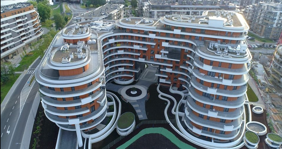 Widok z góry na nowoczesny, zaokrąglony budynek mieszkalny z balkonami i systemem wentylacyjnym na dachu. Widoczne elementy instalacji na dachu budynku.